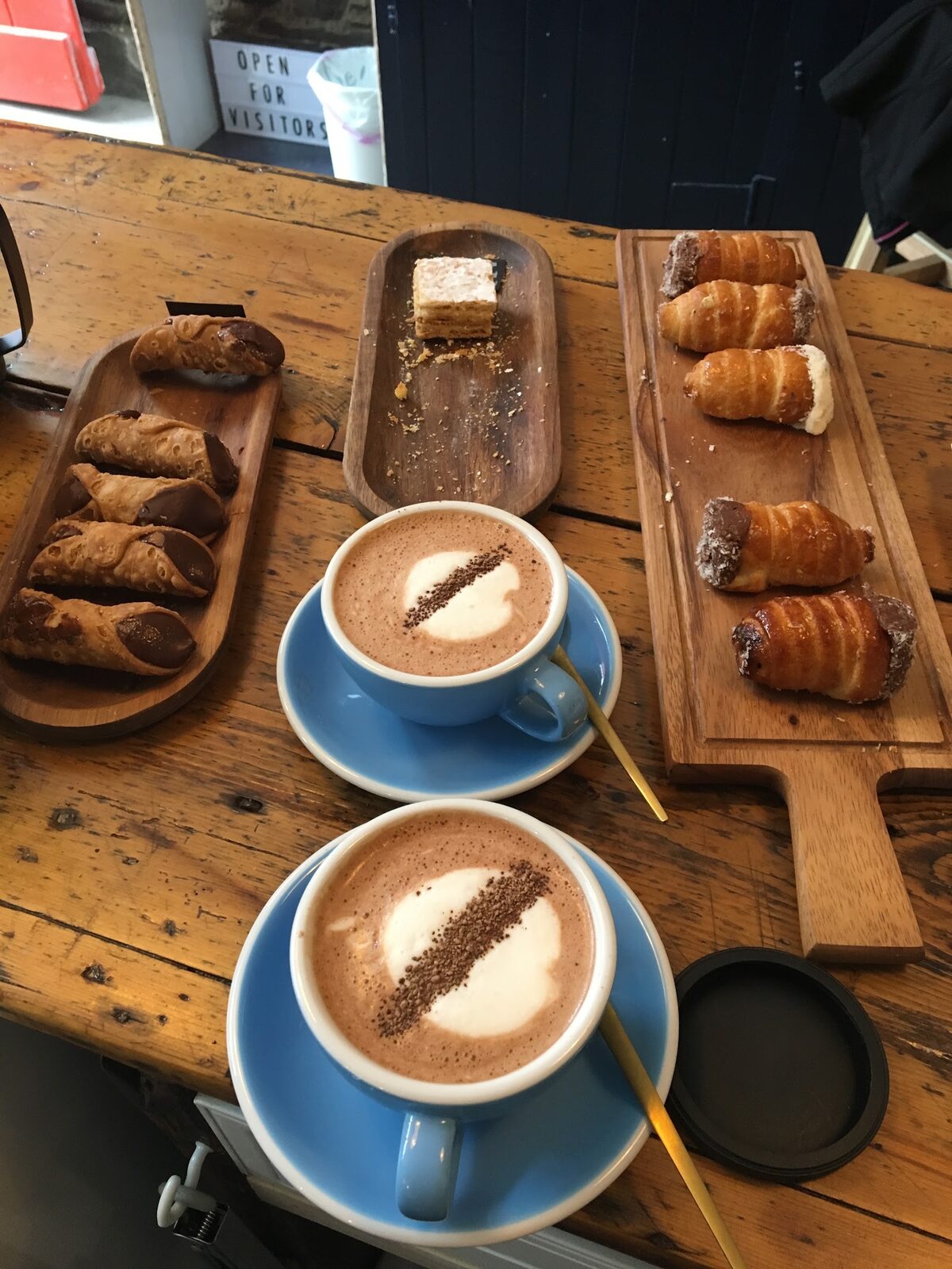 Two hot chocolates in blue cups with cannoli, mille-feuille and cream-filled pastries on a worn wooden counter